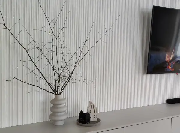 Wazon z gałązkami i lampkami, obok ceramiczne domki. Za telewizorem lamele na ścianie tv tworzą nowoczesne tło.