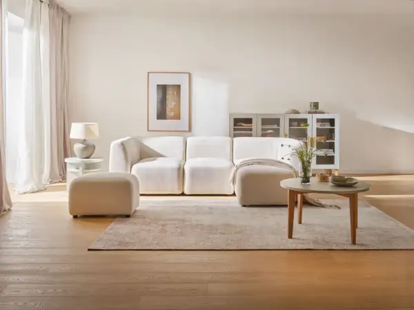 Przytulny salon z kanapą i pufą w beżowym kolorze ścian. Jasne zasłony, dywan i drewniana podłoga tworzą ciepłą atmosferę.
